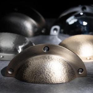 May include: A selection of hammered metal cabinet pulls in bronze, silver, and gold finishes. The semi-circular pulls have a textured surface and two or three screw holes. The image shows a variety of finishes for home decor.
