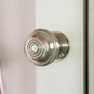 May include: A polished, silver door knob with a circular design. The knob is fixed to a white door. The door knob is round with concentric circles and a small screw.