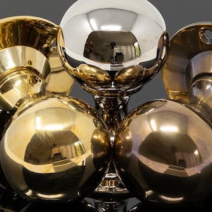 May include: An assortment of polished door knobs and hardware in gold and silver. The central feature is a silver sphere, surrounded by several gold-coloured knobs. The reflective surfaces create a dynamic interplay of light and shadow, enhancing the metallic sheen.