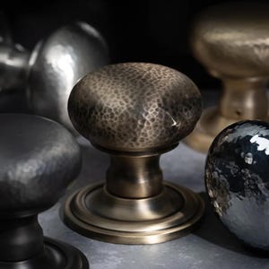 May include: A selection of metal door knobs in different finishes. The knobs vary in shape and texture, with hammered and smooth surfaces. Colours include black, silver, bronze, and gold. Measurements are not available.