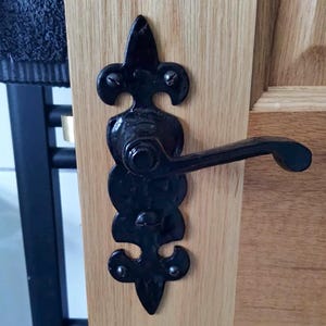 May include: Black metal door handle with an ornate backplate, mounted on a light-coloured wooden door. The handle has a curved lever and is attached with small rivets. The door is made of wood.