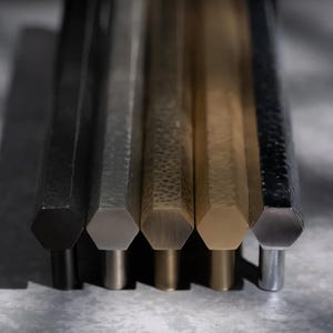 May include: Close-up of five hexagonal cabinet pulls in varying metallic finishes: black, silver, bronze, gold, and brushed nickel. The pulls are arranged vertically, showcasing their geometric design and textured surfaces.