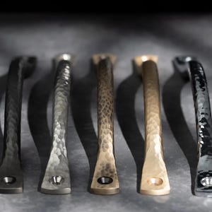 May include: Five metal cabinet pulls in different finishes: black, silver, bronze, gold, and dark grey. Each pull has a hammered texture and a rectangular base with a single screw hole. The pulls are arranged in a row, casting shadows.