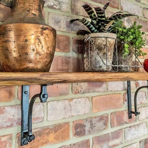 May include: A wooden shelf with black metal brackets fixed to a brick wall. The shelf holds a large copper jug, potted plants in wire baskets, and a small red glass candle holder. The shelf is a warm, natural wood tone, and is approximately 100cm in length.