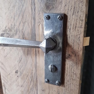 May include: A metal door handle with a rectangular backplate and a geometric handle, mounted on a wooden door. The handle has a textured, aged appearance. The door is made of wood with visible grain, suggesting a vintage or antique style.
