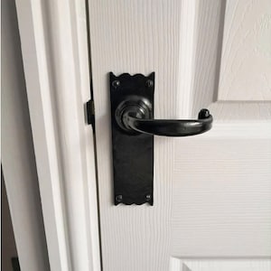 May include: Black door handle with a decorative backplate, mounted on a white wooden door. The handle has a curved design and a matte finish. The backplate has a scalloped edge.
