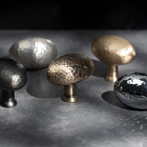 May include: Five hammered metal cabinet knobs in black, silver, bronze, and gold finishes. Each knob has a textured surface and an oval shape. The knobs are displayed on a grey surface.