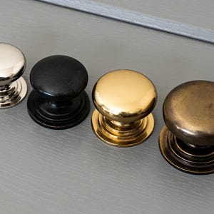 May include: Four cabinet knobs in various finishes: silver, black, gold, and antique brass. Each knob features a rounded top and a circular base with decorative detailing. The knobs are displayed in a row on a grey surface.