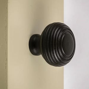 May include: A black, round cabinet knob with a ridged design. The knob is mounted on a cream-coloured cabinet door. The knob's design features concentric circles, creating a textured appearance. The hardware is a classic design.