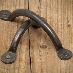 May include: Two dark grey, curved metal handles with mounting holes. The handles are arched and appear to be made of cast iron or a similar material. They are set against a wooden background.