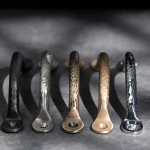 May include: Five metal cabinet pulls in different finishes: dark grey, silver, bronze, gold, and chrome. Each pull has a hammered texture and a curved handle design, suitable for furniture or cabinetry. The pulls are arranged in a row against a neutral background.