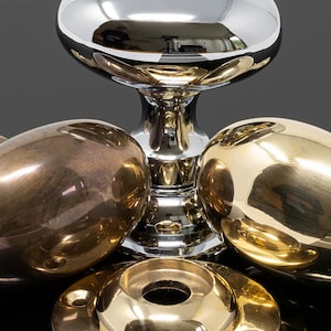 May include: Close-up of various metallic hardware components. The image features a shiny, silver-coloured, decorative knob with a rounded top and a pedestal base. Surrounding it are several gold and bronze-coloured oval-shaped knobs and a circular base.
