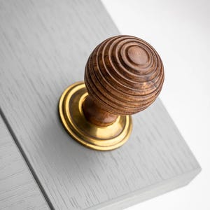 May include: A close-up of a wooden door knob with a brass base. The knob is a sphere with concentric circular grooves. The brass base is circular and has a polished finish. The knob is mounted on a light grey wooden surface.