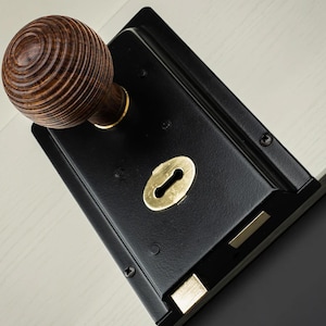 May include: A black door lock with a wooden, spherical knob. The lock has a brass keyhole and accents. The knob features a detailed, concentric ring design. The lock is mounted on a light-coloured surface.