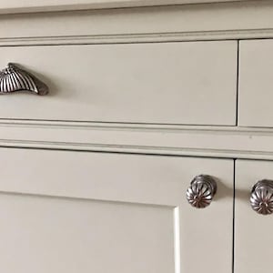 May include: A cream-coloured cabinet with silver hardware. The image shows a drawer with a shell-shaped handle and two doors with floral knobs. The hardware provides an elegant touch to the cabinet's design.