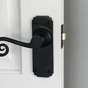 May include: Black iron door handle featuring a decorative scroll design. The handle is mounted on a rectangular backplate with rounded corners and rivets. The door is white, and the latch is gold.