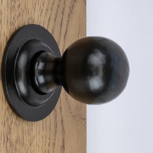 May include: A black, spherical door knob with a textured surface, mounted on a circular backplate. The door knob is attached to a wooden door with visible wood grain.