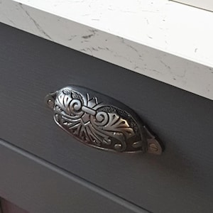 May include: A decorative silver drawer pull with an intricate floral pattern. It's fixed to a dark grey cabinet door, with a white countertop visible above. The pull's design is classic and elegant.