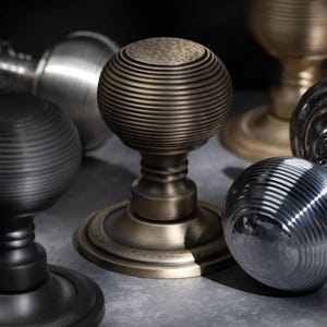 May include: A selection of door knobs in different metallic finishes. The knobs have a round, ribbed design, with finishes including bronze, silver, and black. The knobs are displayed on a neutral surface, highlighting their detailed craftsmanship.