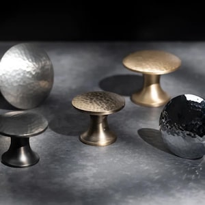 May include: A selection of five metal cabinet knobs in different styles and finishes. The knobs include a large, hammered silver knob, a small, dark grey knob, two bronze knobs, and a hammered silver knob with a rounded shape. The knobs are displayed on a grey surface.