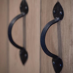 May include: Close-up of two black, curved cabinet handles with a leaf-shaped design at each end. The handles are mounted on light-coloured wooden cabinet doors, showcasing a rustic aesthetic. The handles are made of metal.
