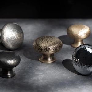 May include: Five hammered metal cabinet knobs in silver, bronze, gold, and black. Each knob has a rounded, textured surface and a stem for mounting. The knobs are displayed on a grey surface, highlighting their different colours and textures.