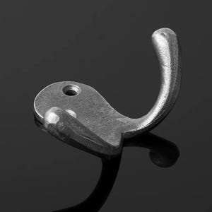 May include: A single, antique-style metal hook in a grayscale image. The hook has a curved design with a rounded top and a small, flat base with a single hole for mounting. The metal has a slightly textured, aged appearance.