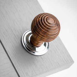 May include: A close-up shot of a wooden cabinet knob with a grooved, spherical design. The knob is mounted on a silver base and attached to a light grey cabinet door. The wood has a warm brown tone.