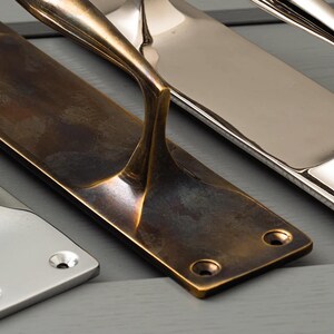 May include: Close-up of three metal door pulls in different finishes: antique brass, polished nickel, and chrome. The pulls have a rectangular base with mounting holes and a curved handle. The antique brass pull shows signs of wear.