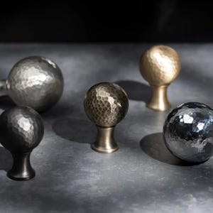 May include: Five decorative cabinet knobs with hammered metal finishes. The knobs are in various colours, including silver, gold, and black. Each knob has a round, textured sphere design.