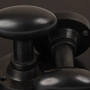 May include: Two black, oval door knobs with a matte finish. They are mounted on circular backplates. The design is simple and classic, suitable for various interior styles. The hardware is made of metal. The knobs are a dark, muted colour.