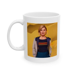 May include: White ceramic mug with a colorful image of a woman with short blonde hair wearing a blue and grey jacket and blue pants against a yellow background.