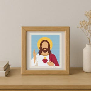 May include: A framed cross-stitch art piece depicting a figure with a halo, long brown hair, and a beard. The figure wears a white robe and a red garment, with a heart symbol on the chest. The background is light blue, and the frame is made of wood.