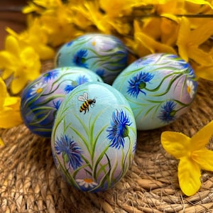 May include: Four hand-painted Easter eggs with a light blue background, adorned with blue cornflowers, green stems, and a bee. The eggs are arranged on a woven basket, surrounded by yellow flowers.