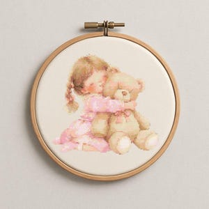 以下が含まれることがあります： ピンクのドレスを着た女の子がテディベアを抱きしめているクロスステッチ刺繍枠。女の子は三つ編みの茶色の髪をしています。テディベアは薄茶色です。刺繍は木製の枠にセットされています。