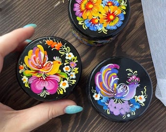 May include: Three black, round boxes with lids, each adorned with vibrant floral and bird designs. The hand-painted artwork features bright colors like yellow, pink, and blue. The boxes sit on a dark wooden surface.