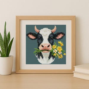 Pode incluir: Um ponto de cruz emoldurado de uma vaca com manchas pretas e brancas, segurando flores amarelas e brancas na boca. A vaca tem chifres e orelhas cor-de-rosa. O fundo é verde-azulado.