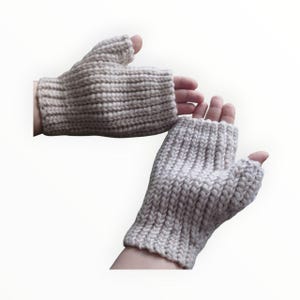 May include: A pair of light beige fingerless gloves, knitted with a thick, textured yarn. The gloves cover the hand up to the knuckles, leaving the fingers free. The knitted pattern is visible, with vertical ribbing. The gloves are shown on a white background.
