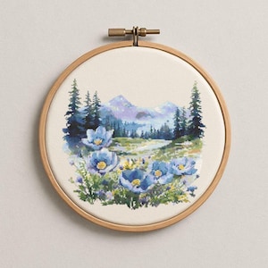 以下が含まれることがあります： 木製のフレームに入ったクロスステッチ刺繍は、穏やかな山の風景を描いています。 前景には青い花、川、常緑樹が描かれています。 山は柔らかな紫の色合いで、空は水色です。