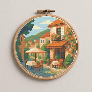 May include: A cross-stitch embroidery of a European village scene, set in a wooden hoop. The artwork depicts a building with an orange tile roof, surrounded by lush greenery and a cafe with tables and an umbrella. The sky is blue with fluffy white clouds.