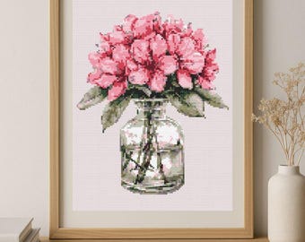 May include: Cross-stitch artwork of pink flowers in a clear glass vase. The framed piece is set against a light pink background, with a wooden frame. The artwork is displayed on a wooden surface.