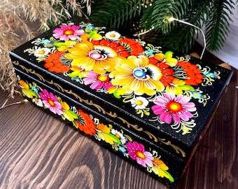 May include: A black wooden box with a vibrant floral design. The flowers are painted in bright colours including yellow, pink, orange, and white. The box is rectangular and has a hinged lid.