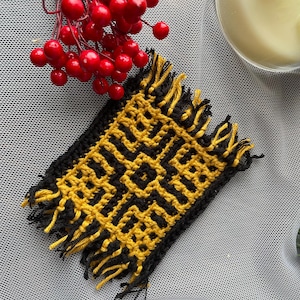 May include: A square, handmade coaster with a geometric pattern in black and yellow. The coaster has a fringed edge and is placed on a textured, light gray surface. Red berries and a candle are in the background.