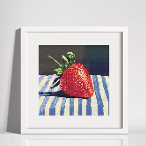 Op de afbeelding: Een ingelijst kruissteekkunstwerk met een helderrode aardbei met groene bladeren. De aardbei ligt op een blauw-wit gestreept oppervlak. De achtergrond is een donkere, effen kleur.