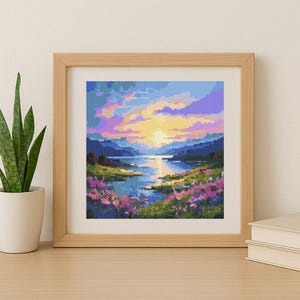 Pode incluir: Uma paisagem em ponto cruzada emoldurada retrata um pôr do sol sobre um lago, com flores rosa e roxas em primeiro plano. O céu transita do amarelo para o azul e roxo. A moldura é castanho claro.