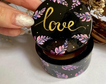 May include: A round, black wooden box with a lid. The lid and sides are decorated with pink and purple floral designs and gold accents. The word "love" is written in gold script on the lid.
