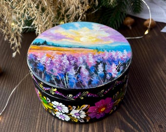 May include: A round, hand-painted wooden box with a lid featuring a landscape of lavender fields under a sunset sky. The box is black with floral designs around the base. The lid's painting includes shades of purple, blue, yellow, and orange.