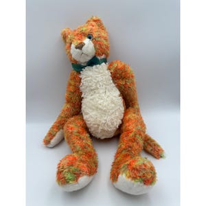 May include: A plush cat toy with orange, green, and white fur. The cat has a white chest, a green bow tie, and a brown nose. The toy is sitting with its legs extended.