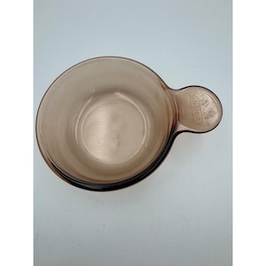 May include: A brown glass Vision saucepan with a handle. The round bowl has a dark rim and a handle with the Vision logo. The glass is transparent, allowing the contents to be seen. The saucepan is designed for cooking.