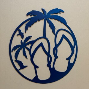May include: A blue metal wall decor featuring a circle with two flip flops and palm trees inside. The flip flops are in the foreground and the palm trees are in the background.